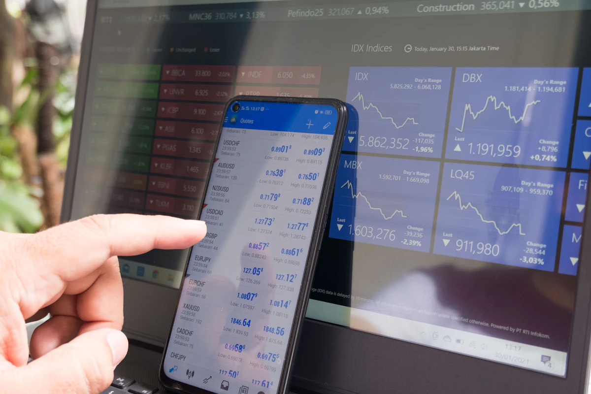 A finger pointing at a trading app on a phone in front of a laptop